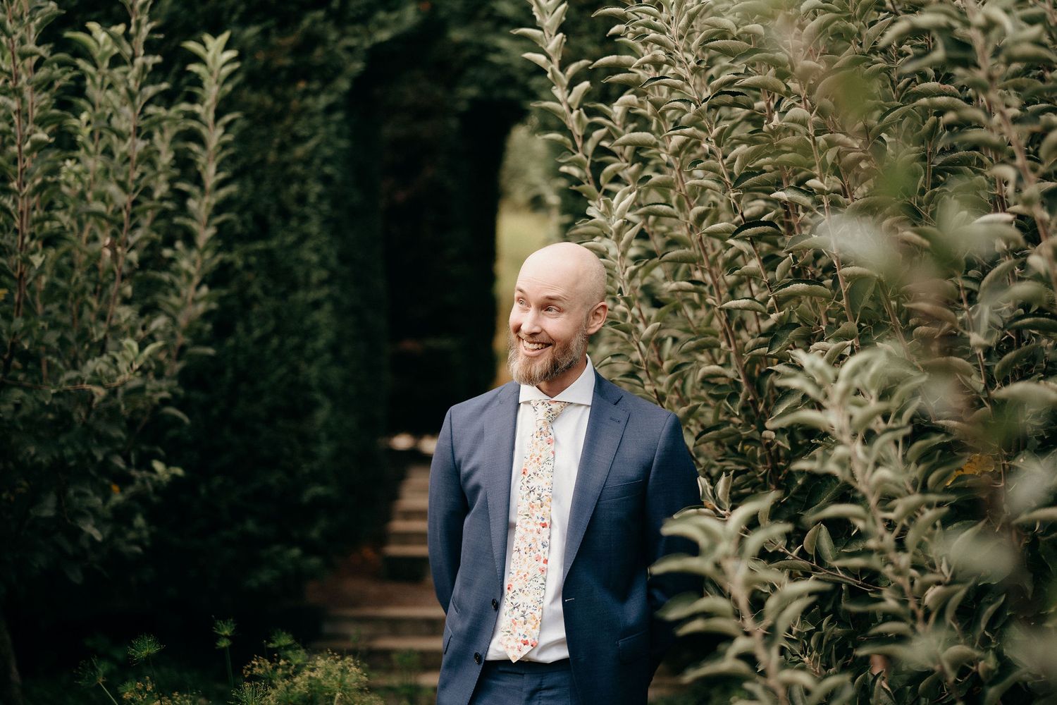 Someone in a navy blue suit and patterned shirt smiles while standing among tall green plants in a garden setting.