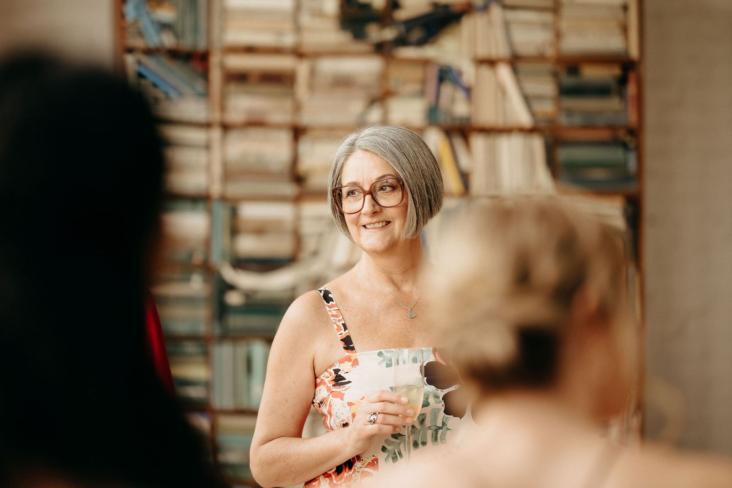 A guest at a social gathering smiles while holding a drink cup in a warmly lit room with bookshelves in the background.