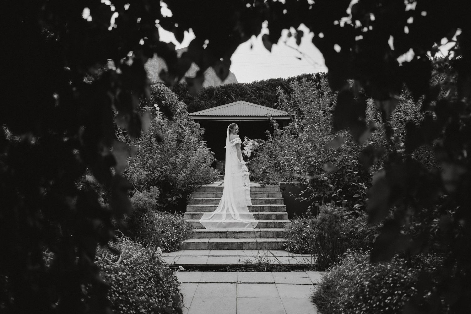 A solitary figure stands at the top of stone garden steps under a white umbrella in a black and white photograph.