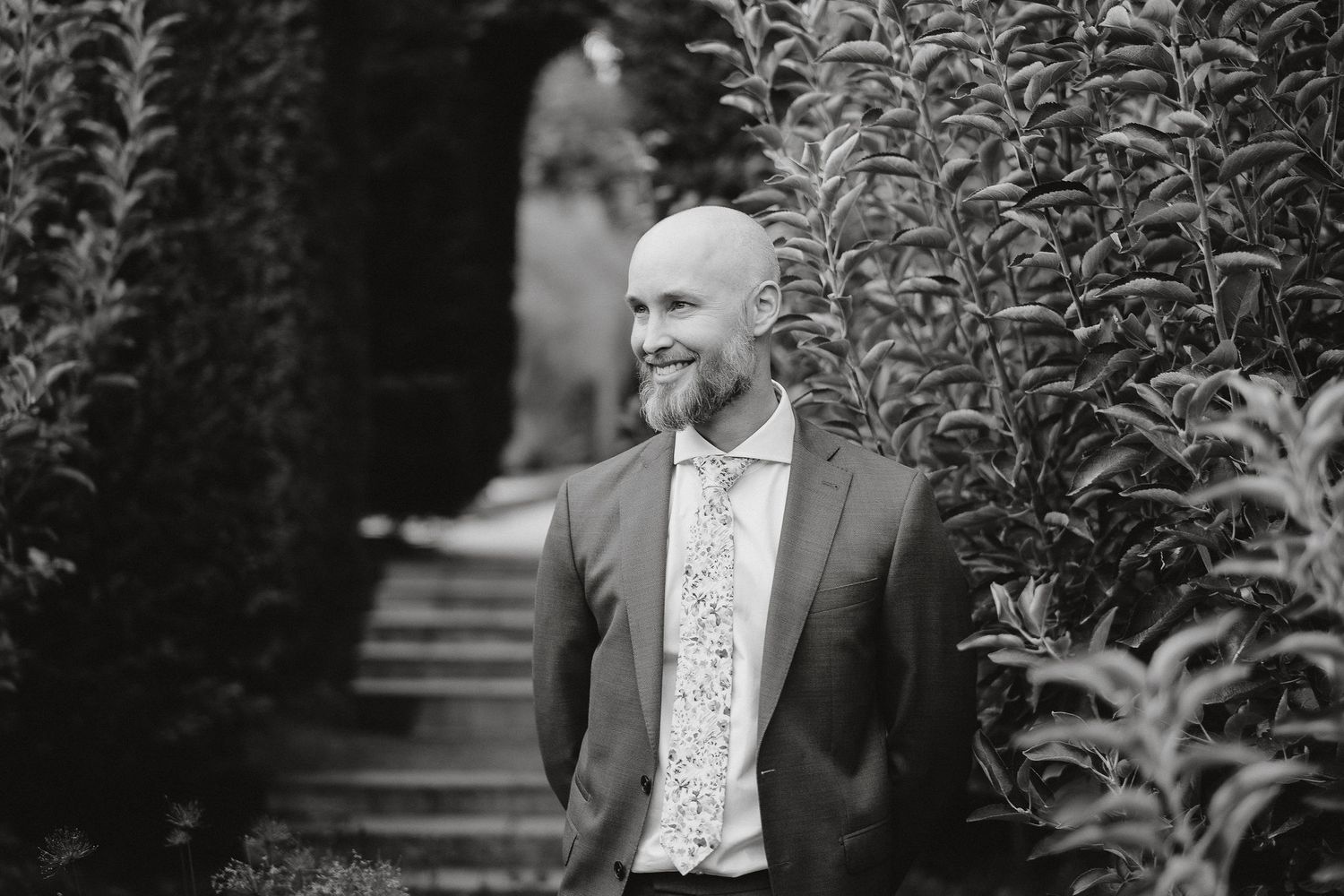 A black and white portrait of a smiling person in a suit jacket and floral shirt standing among lush garden foliage.