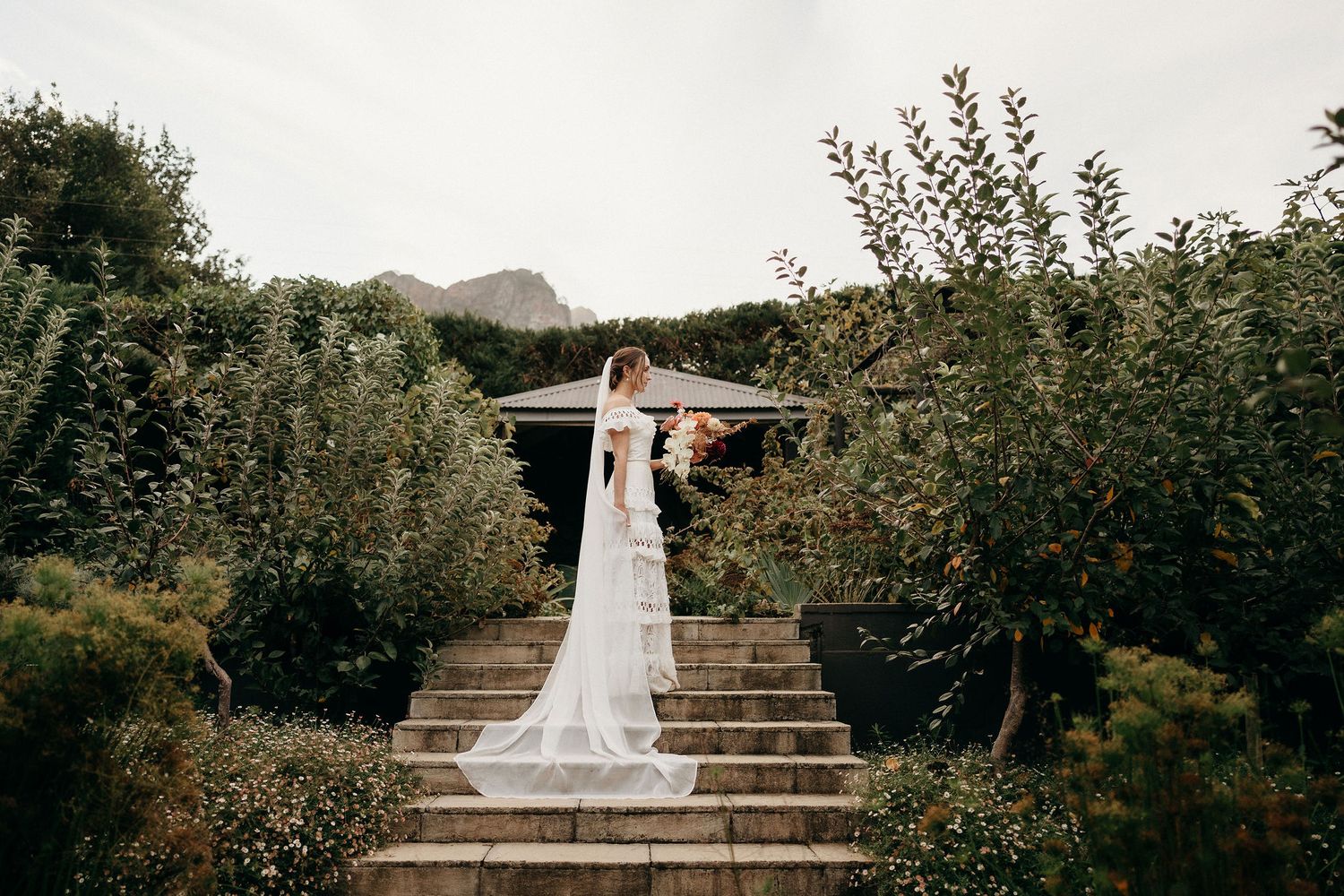 A scenic wedding photo taken on stone steps surrounded by lush gardens and greenery on a cloudy day.