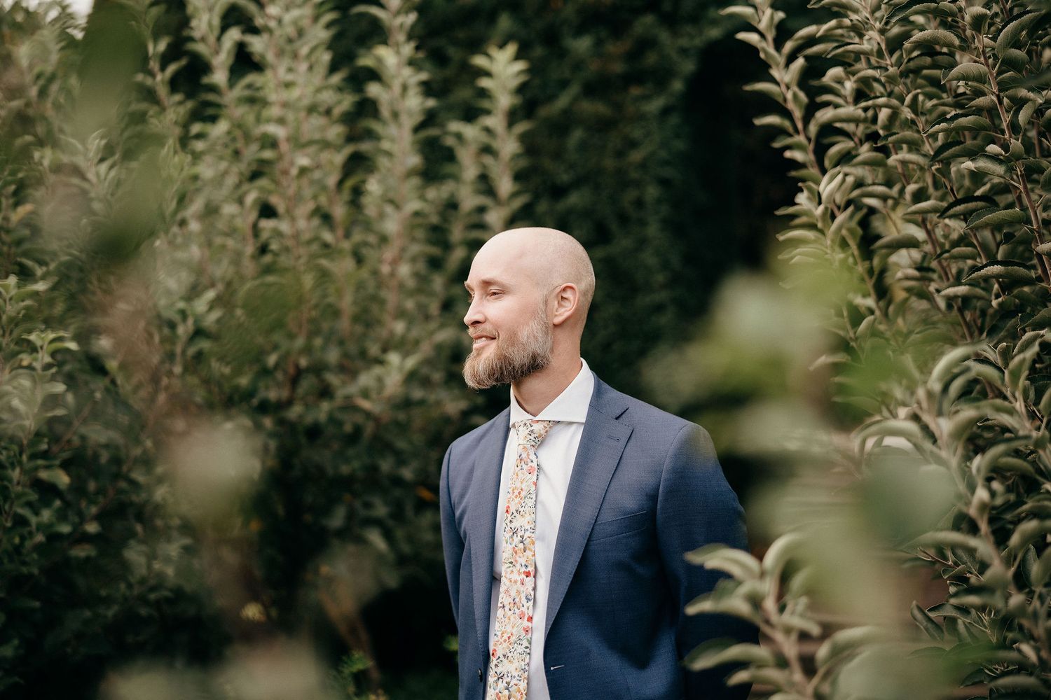 A person in a navy suit and floral tie stands among green foliage in a garden setting, looking to the side.