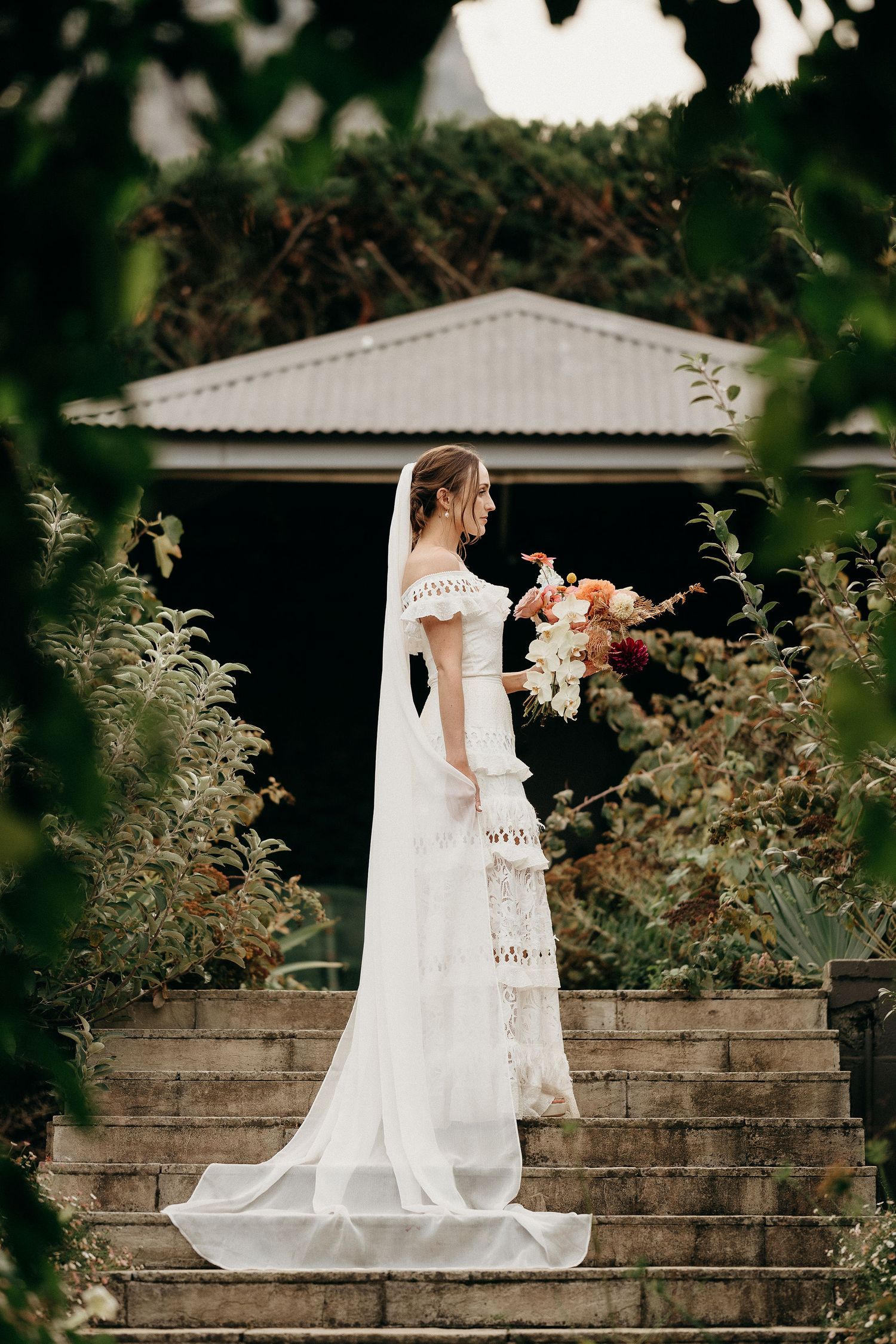 A bride in a lace off-shoulder wedding gown gazes at her bouquet of peach flowers while standing on stone steps.