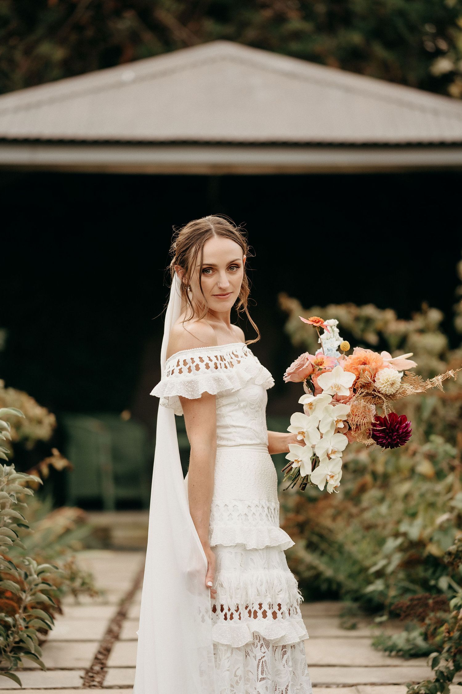 Bride in bohemian off-shoulder lace wedding dress holds peach and white floral bouquet in garden setting.