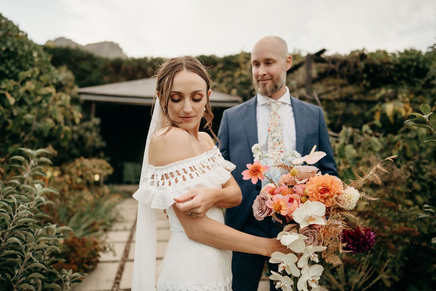 A couple in wedding attire poses outdoors with a vibrant coral and peach floral bouquet against natural greenery.