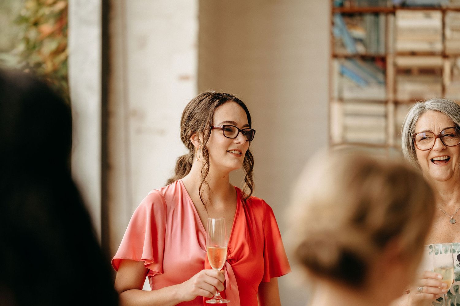 Two guests laugh together at a social event in a bright indoor setting while wearing glasses.