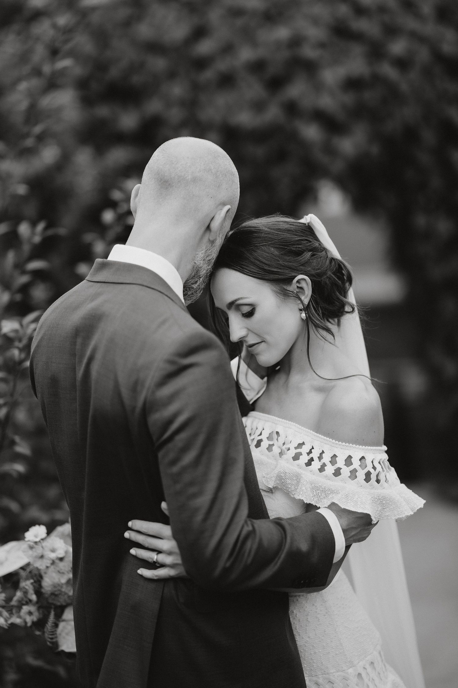 Black and white wedding photo showing intimate moment between bride in lace dress and groom in suit embracing outdoors.