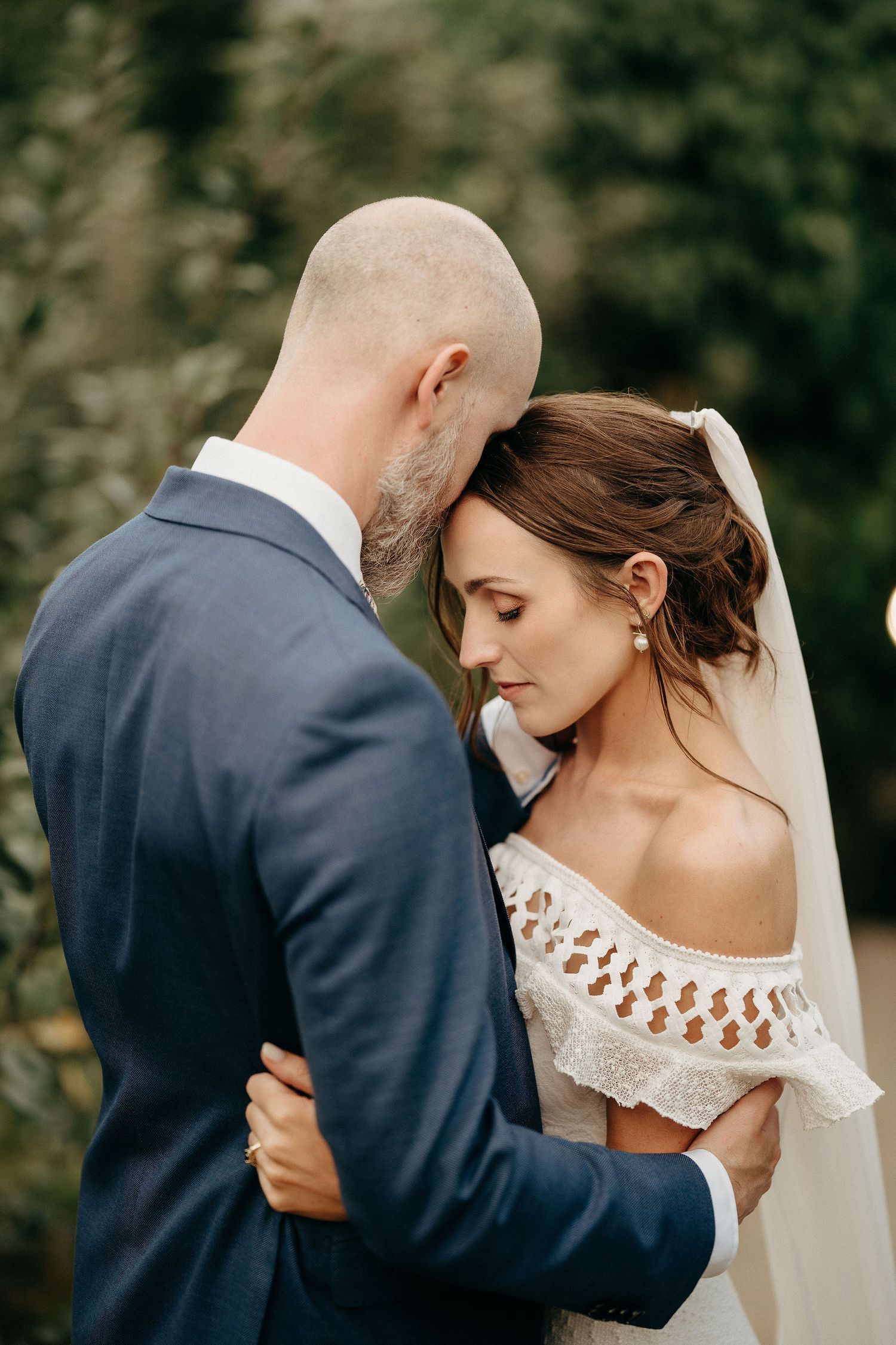 A romantic wedding photo shows a tender embrace between newlyweds in a navy suit and off-shoulder lace dress outdoors.