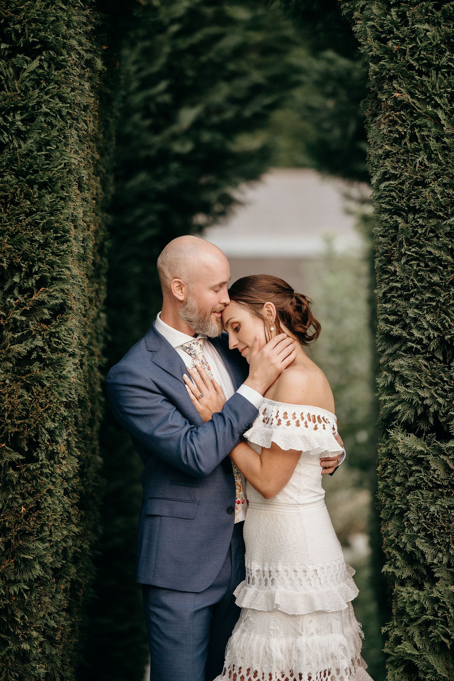 A loving couple shares an intimate moment in a formal garden setting framed by tall evergreen hedges.