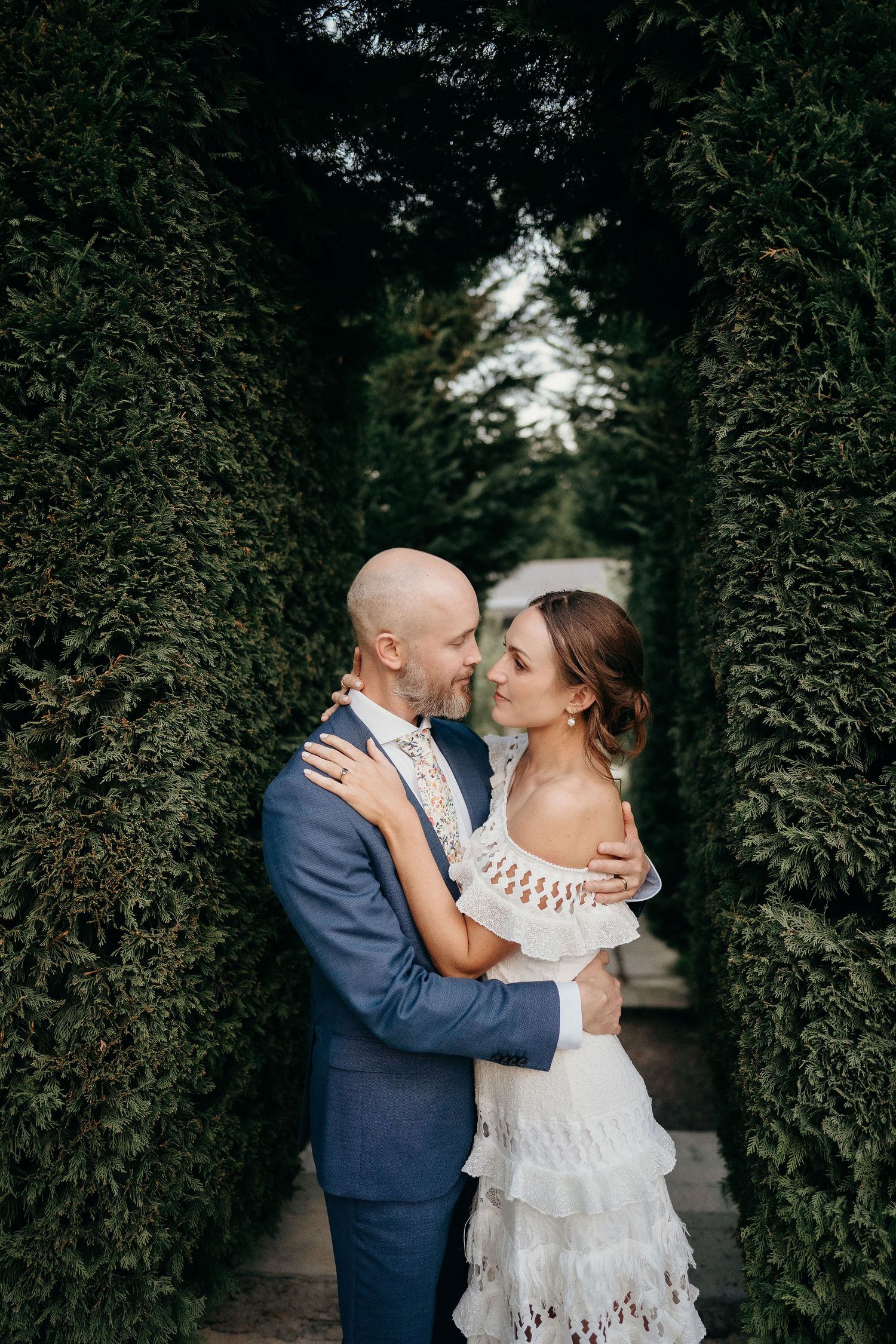Newlyweds share an intimate moment in a green hedge tunnel wearing blue suit and white lace dress.