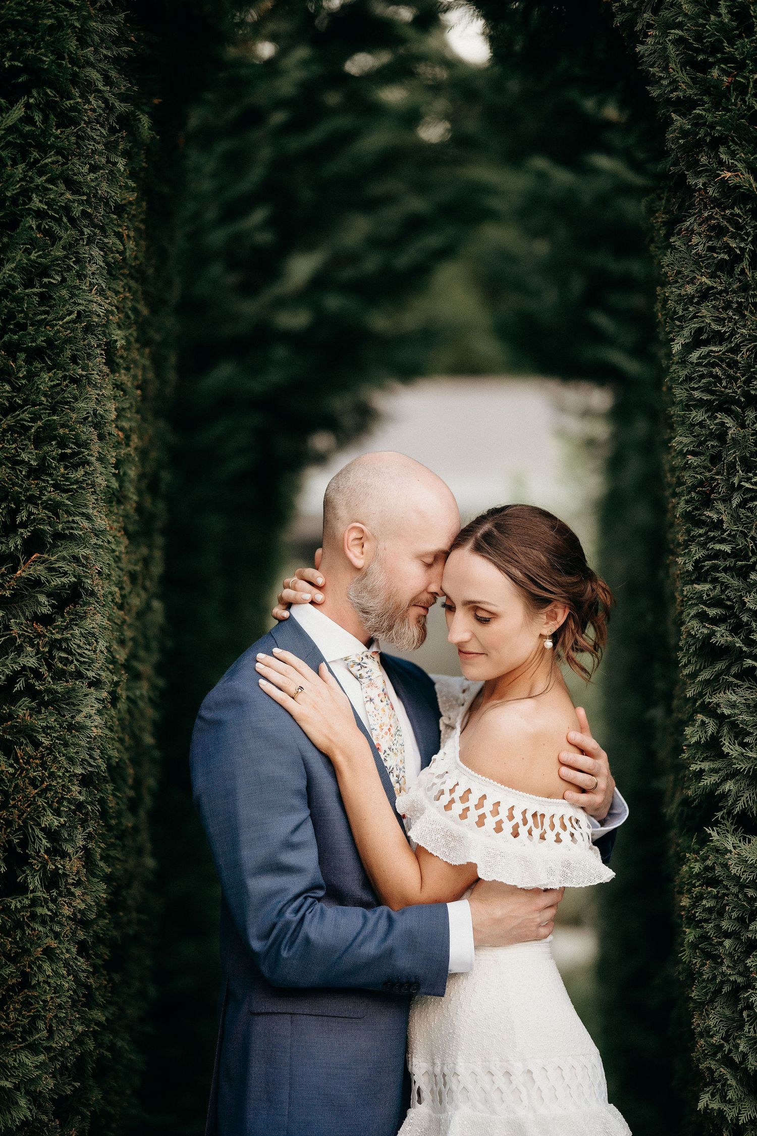 A tender wedding moment captured between a couple in an elegant garden setting with tall hedges.