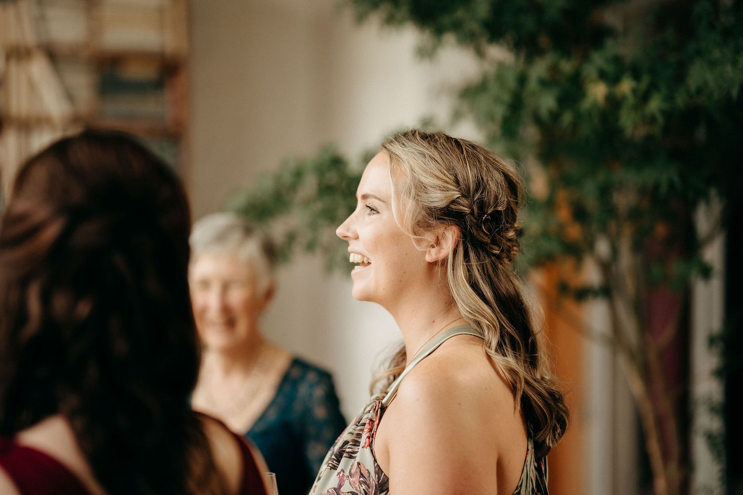 A guest smiles at an indoor social gathering with warm lighting and greenery visible in the background.