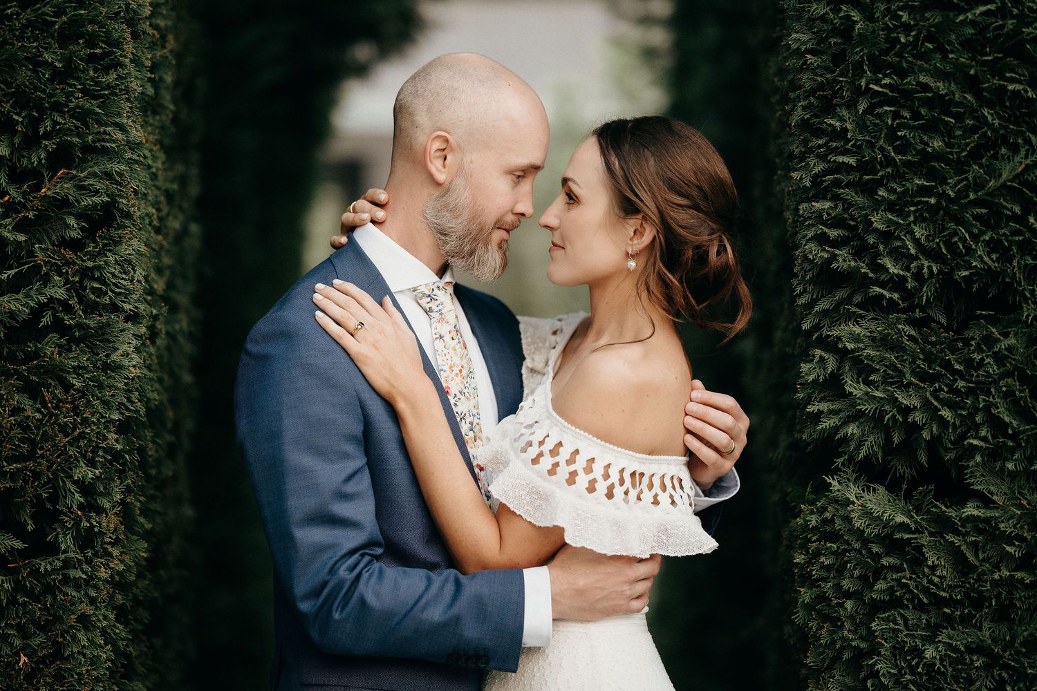A romantic wedding couple embraces between tall green hedges with the bride wearing a white off-shoulder dress.