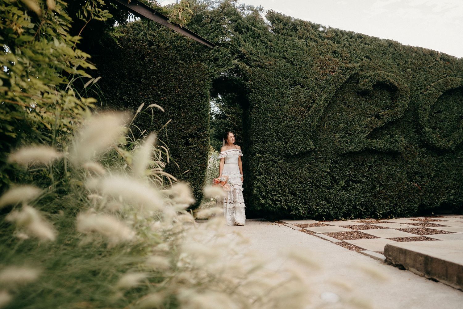 Figure in white dress stands by lush garden hedge wall on stone pathway in romantic outdoor setting.
