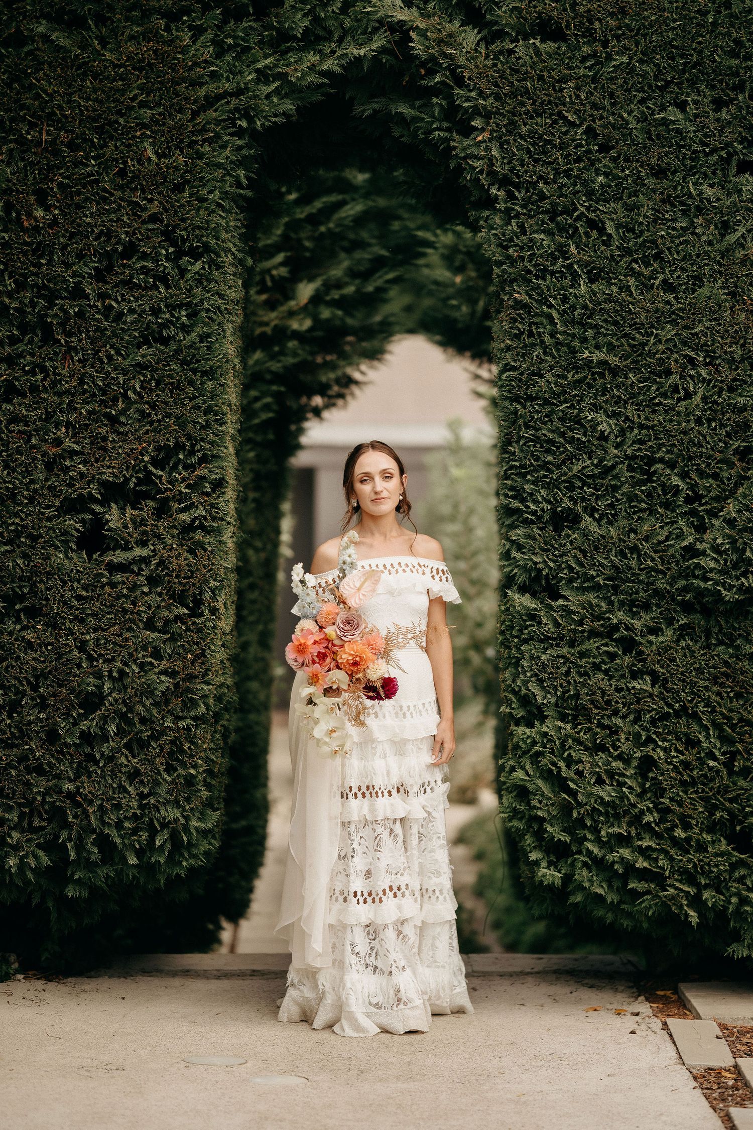 Bride in elegant lace off-shoulder dress stands in garden archway holding vibrant orange and pink floral bouquet.