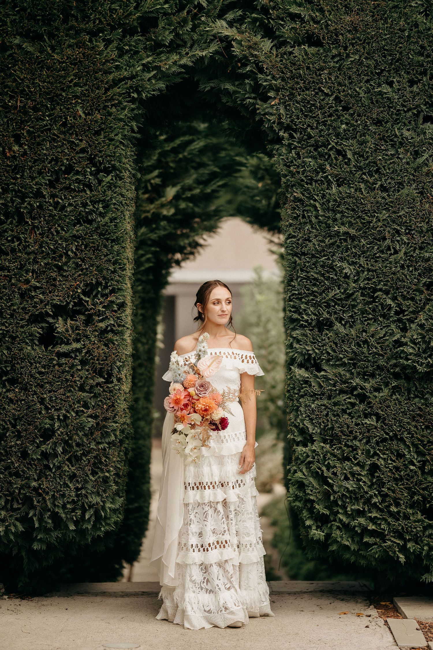A bride in a bohemian lace off-shoulder wedding dress holds a coral bouquet while standing in a garden archway.