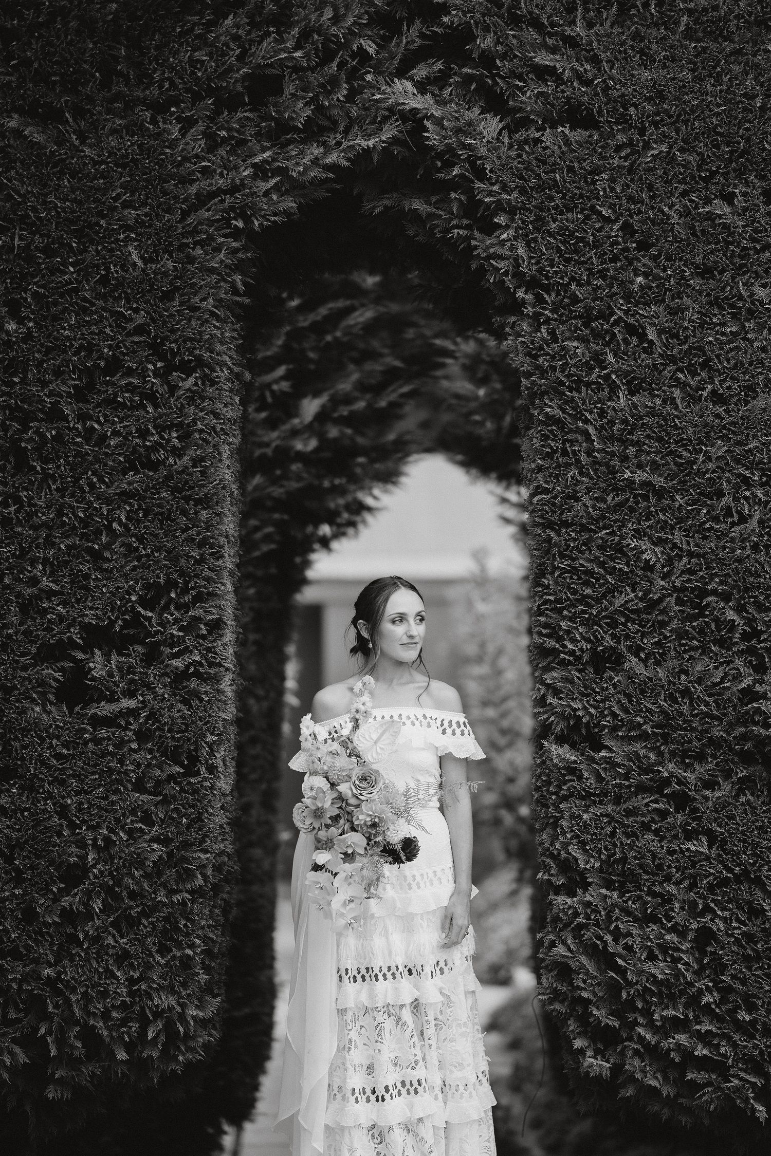 A black and white photo of a bride in a lace wedding dress holding a bouquet, framed by a garden archway.