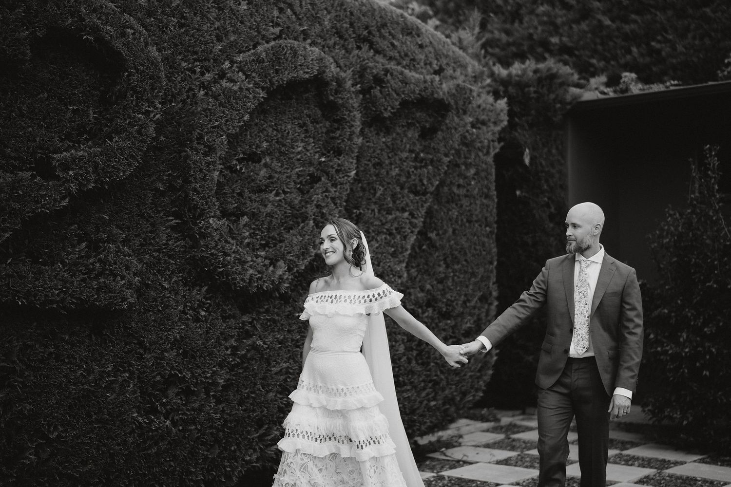 A black and white wedding photo of a couple holding hands in a garden with ivy-covered walls.