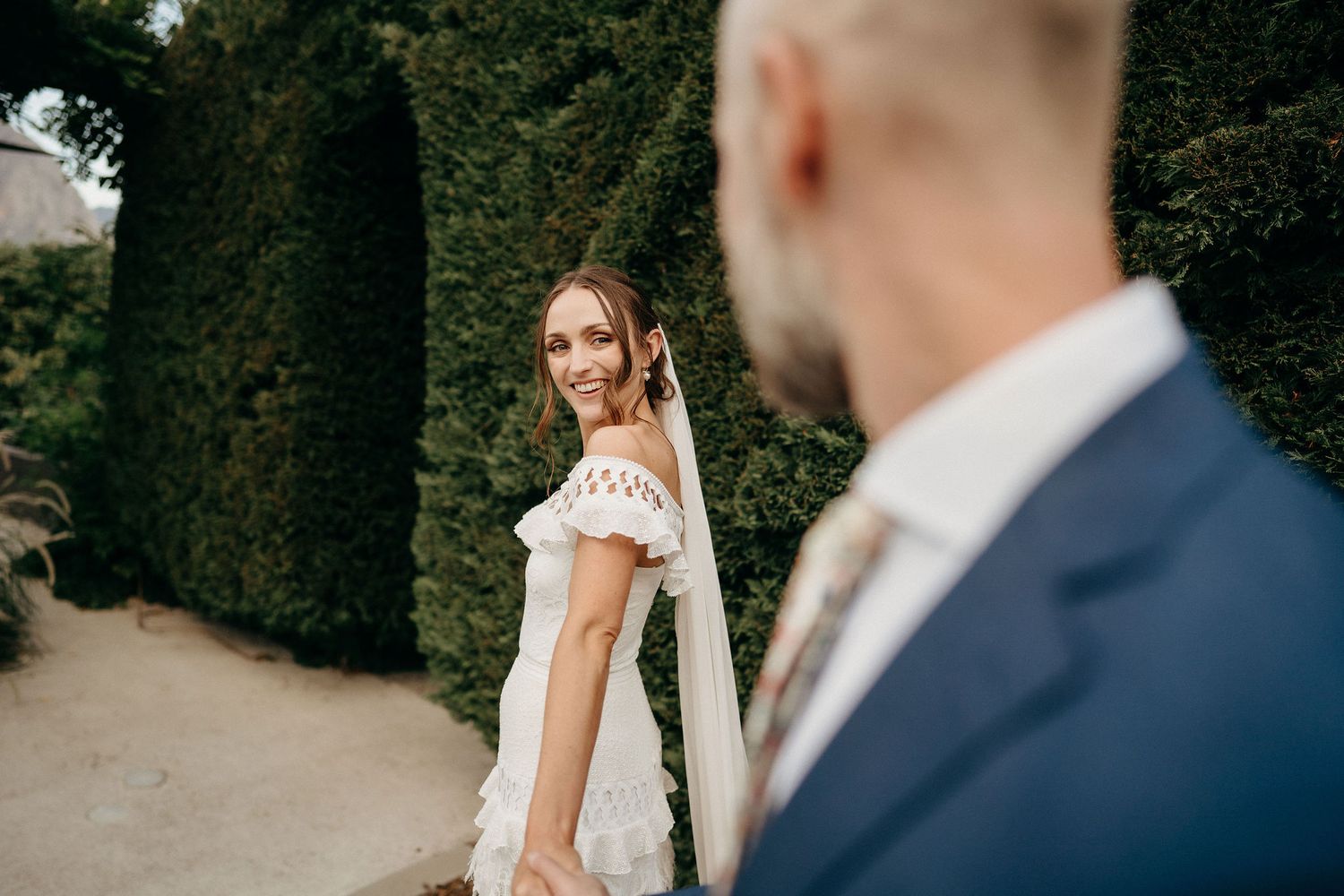 A couple in wedding attire stand together in a garden setting with green hedges.