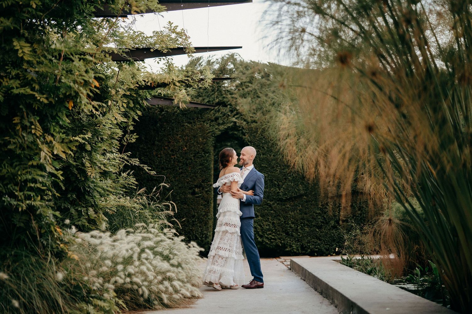A couple embraces on a garden pathway surrounded by lush greenery and ornamental grasses.