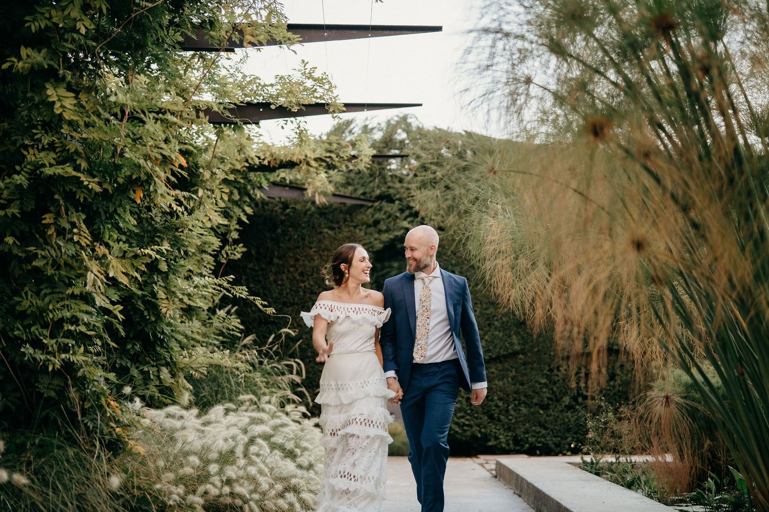 A couple in wedding attire walks hand in hand along a garden path surrounded by lush greenery and tall grasses.