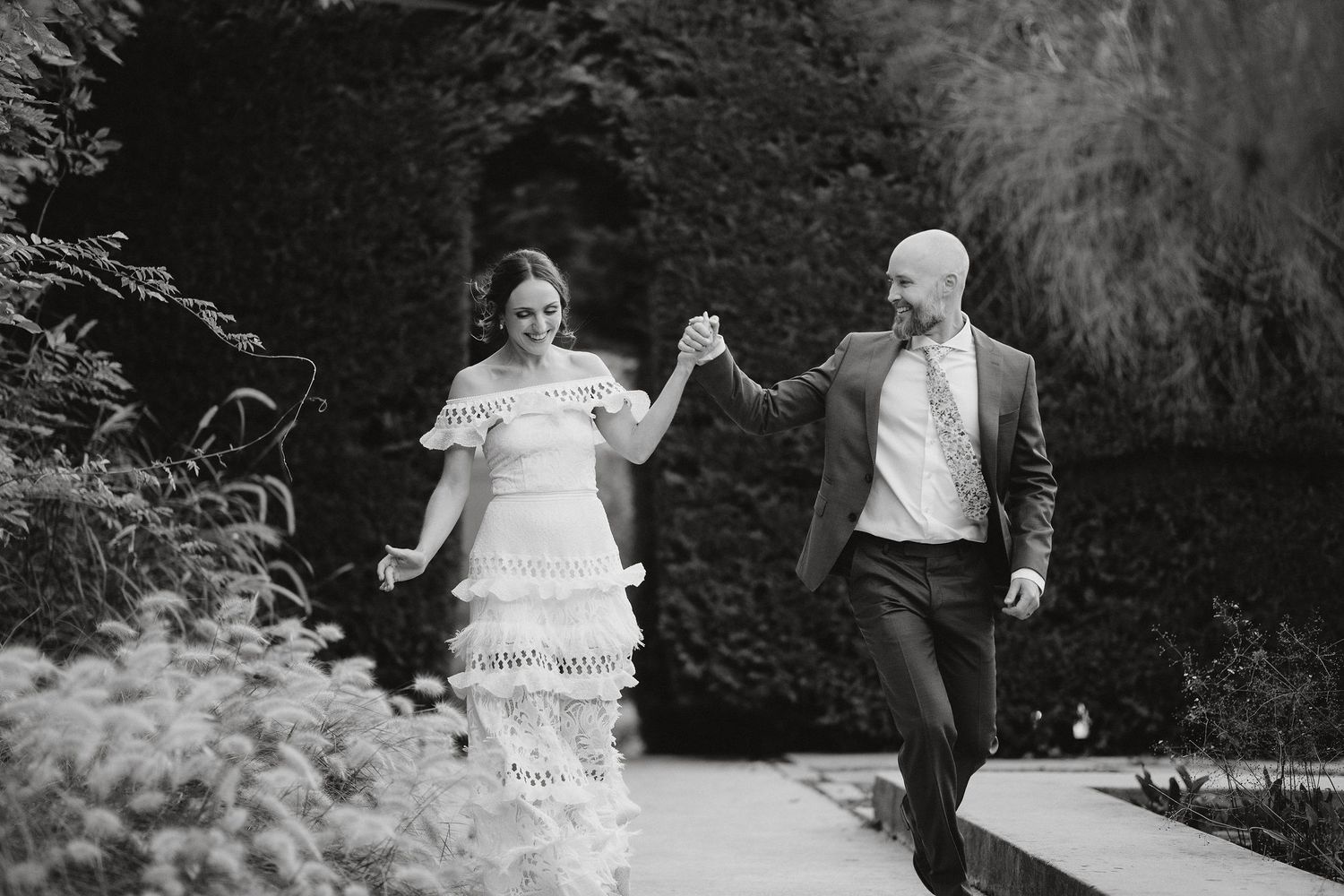 A couple walks joyfully hand in hand along a garden path in black and white wedding attire.