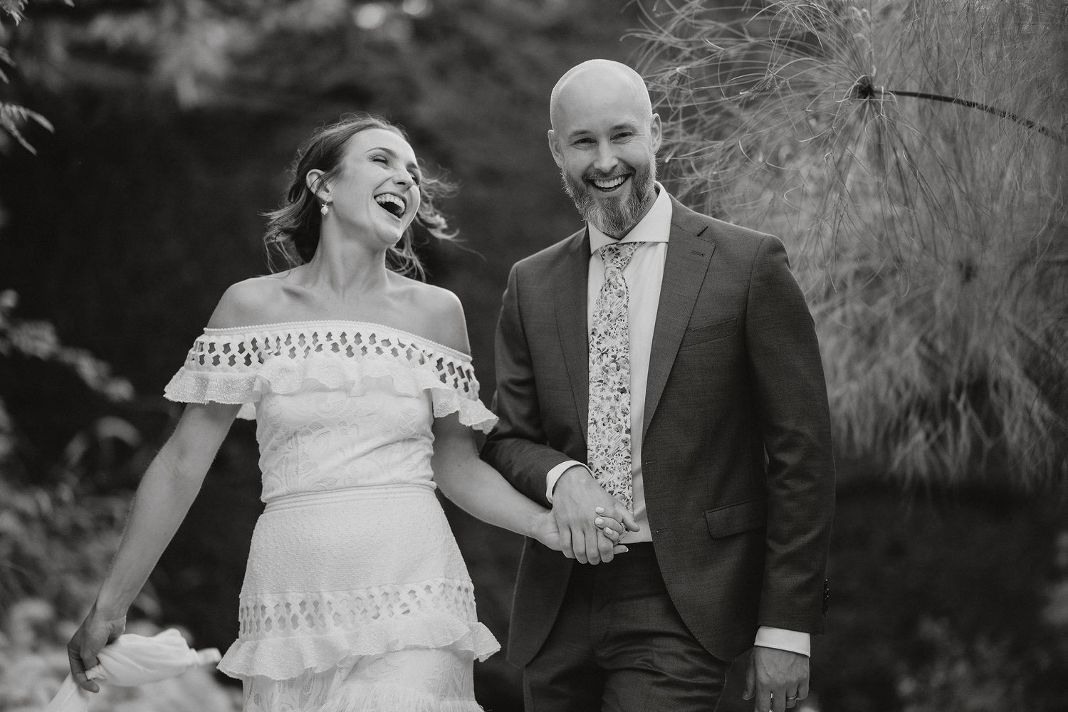 A joyful black and white wedding photo of a couple walking outdoors with the bride wearing a lace off-shoulder dress.