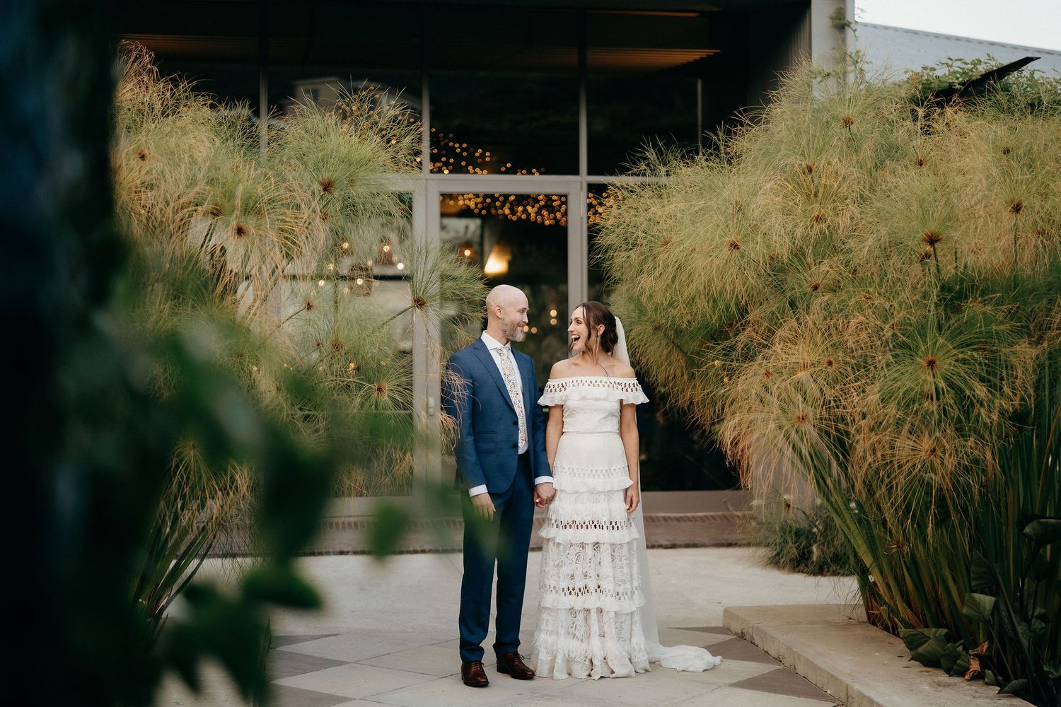 A couple in wedding attire stands on a pathway surrounded by tall ornamental grasses and modern architecture.