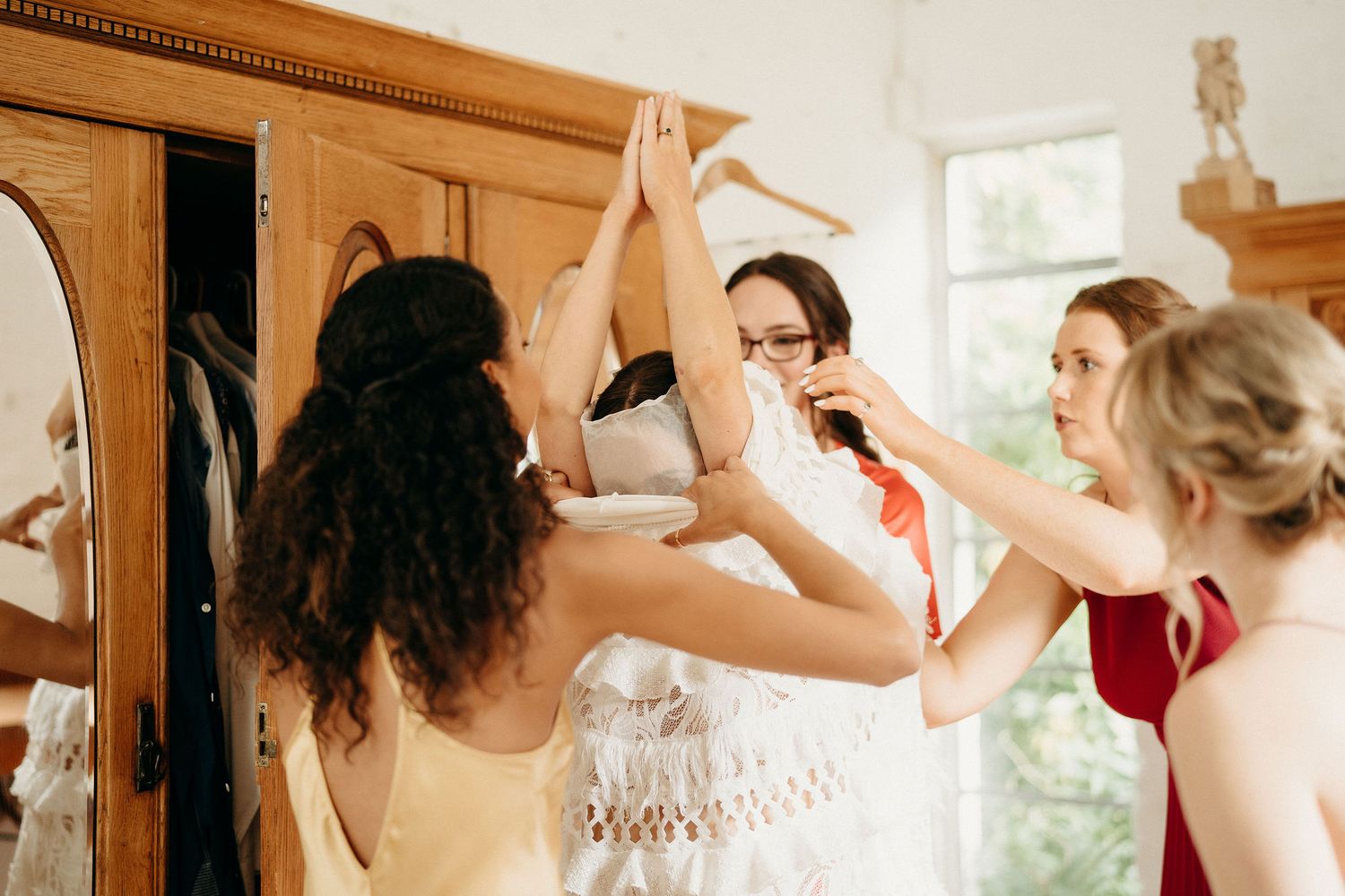 A group of women in formal attire help lift a white wedding dress off a wooden wardrobe.