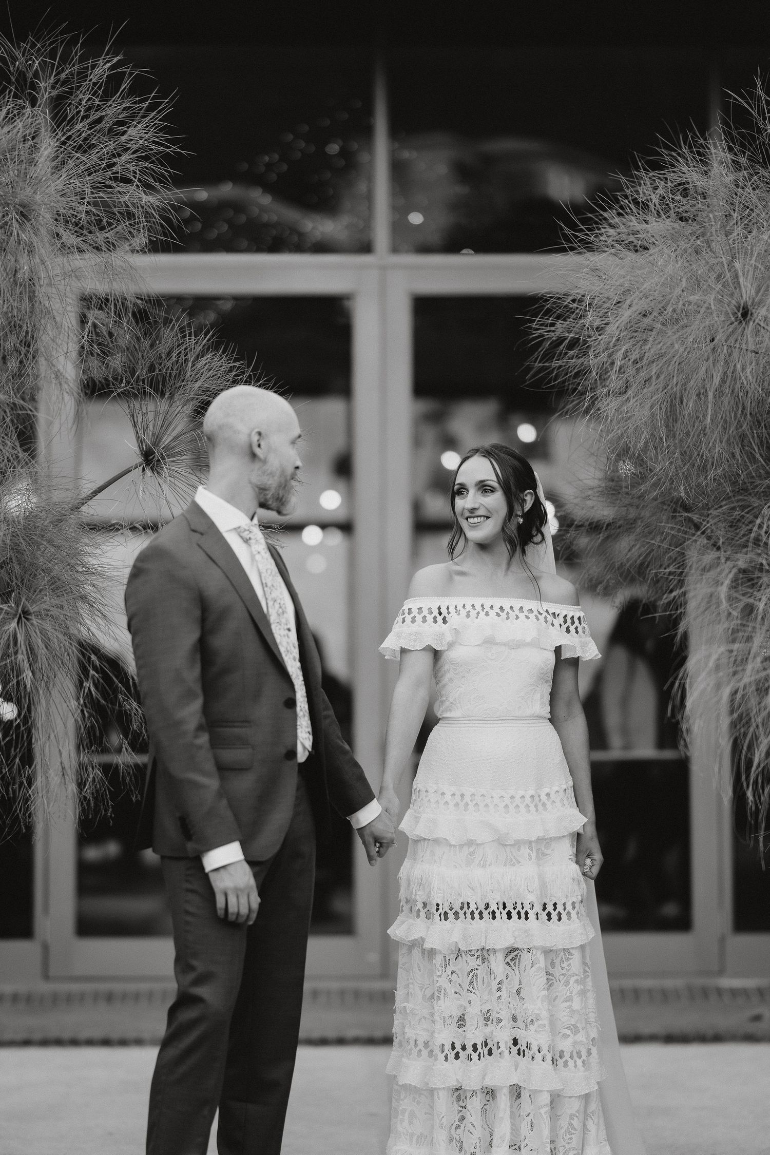 Black and white wedding photo of a couple standing together in front of tall decorative pampas grass and window backdrop.
