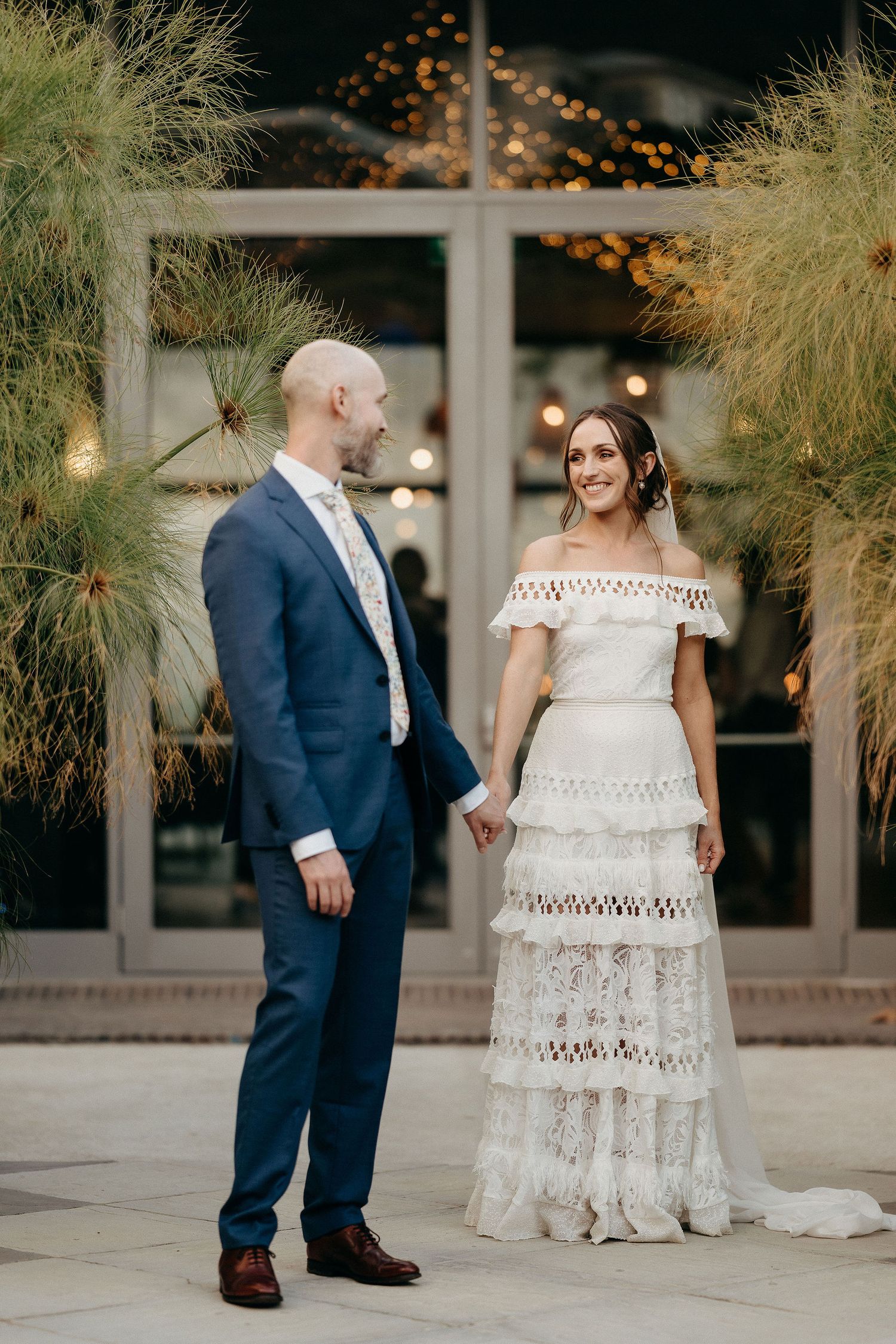 A couple poses at their wedding, with bride in a tiered lace gown and groom in navy suit with twinkle lights behind them.