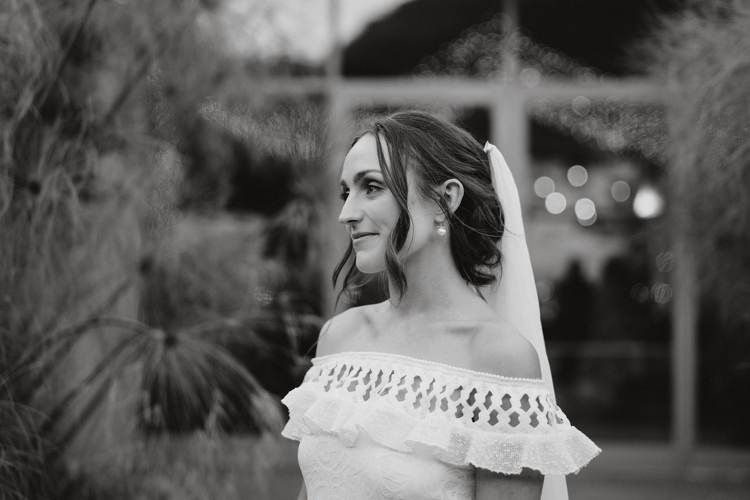 Black and white photo showing an elegant bride in an off-shoulder dress with lace trim gazing to the side during a wedding.