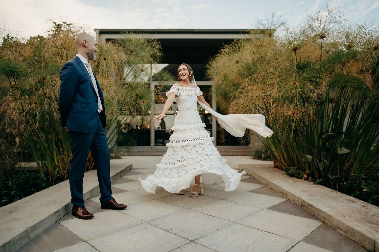 A couple poses outdoors at a garden venue with a flowing white wedding dress catching the breeze.