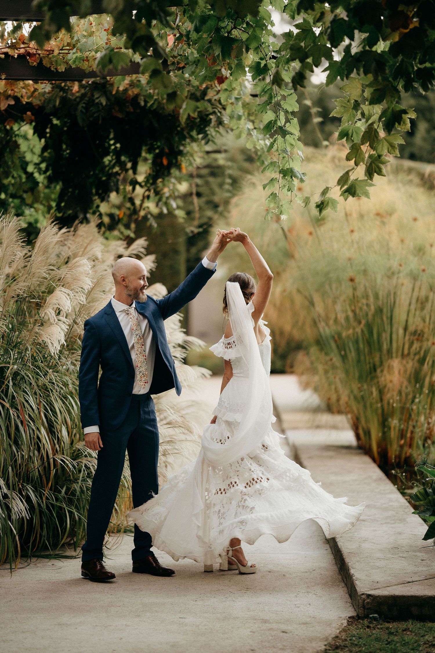 A couple dances together on a garden path with the bride twirling in a flowing white wedding dress among green foliage.