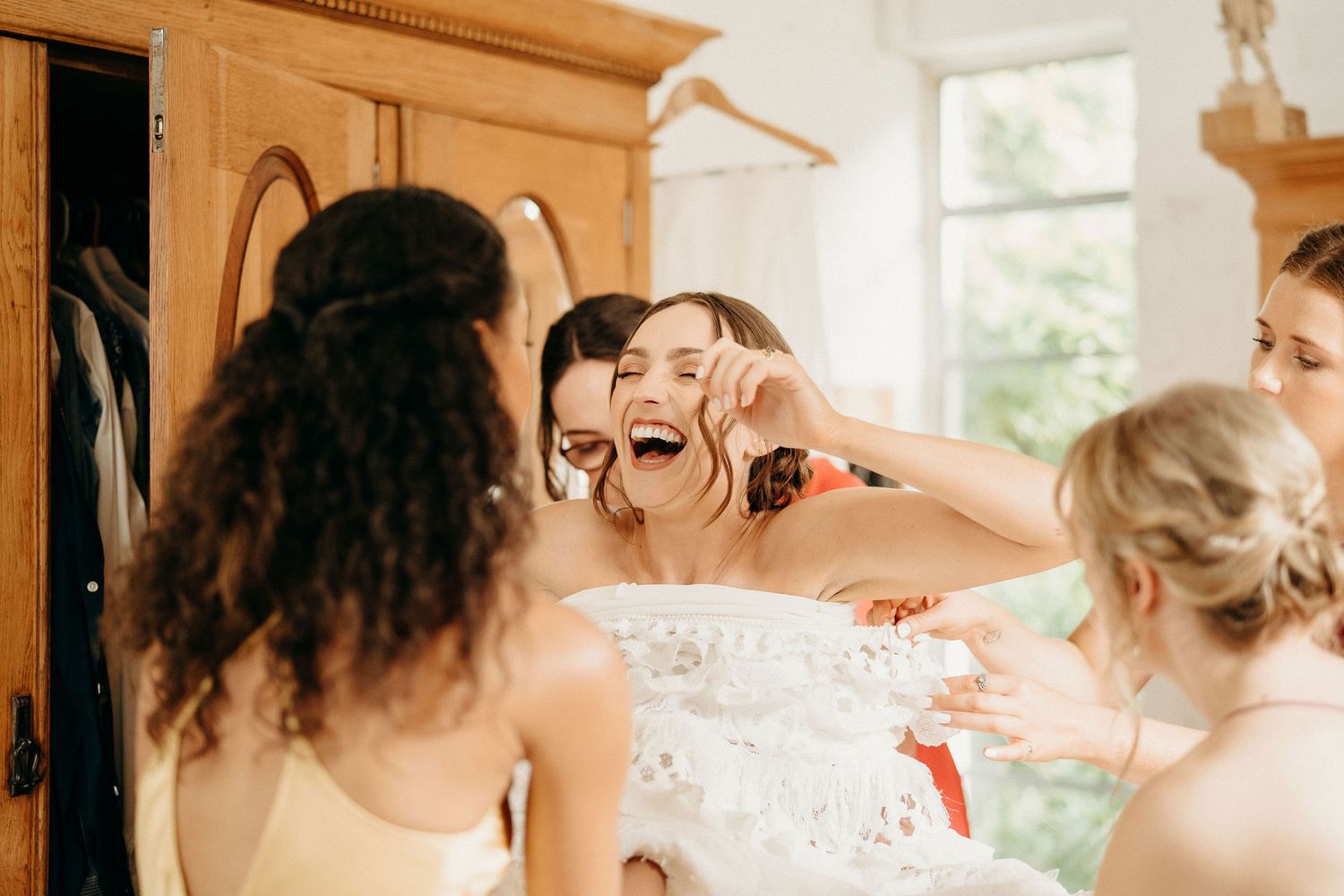 Group shares a joyful moment during wedding preparations in a sun-lit room with warm interior tones.