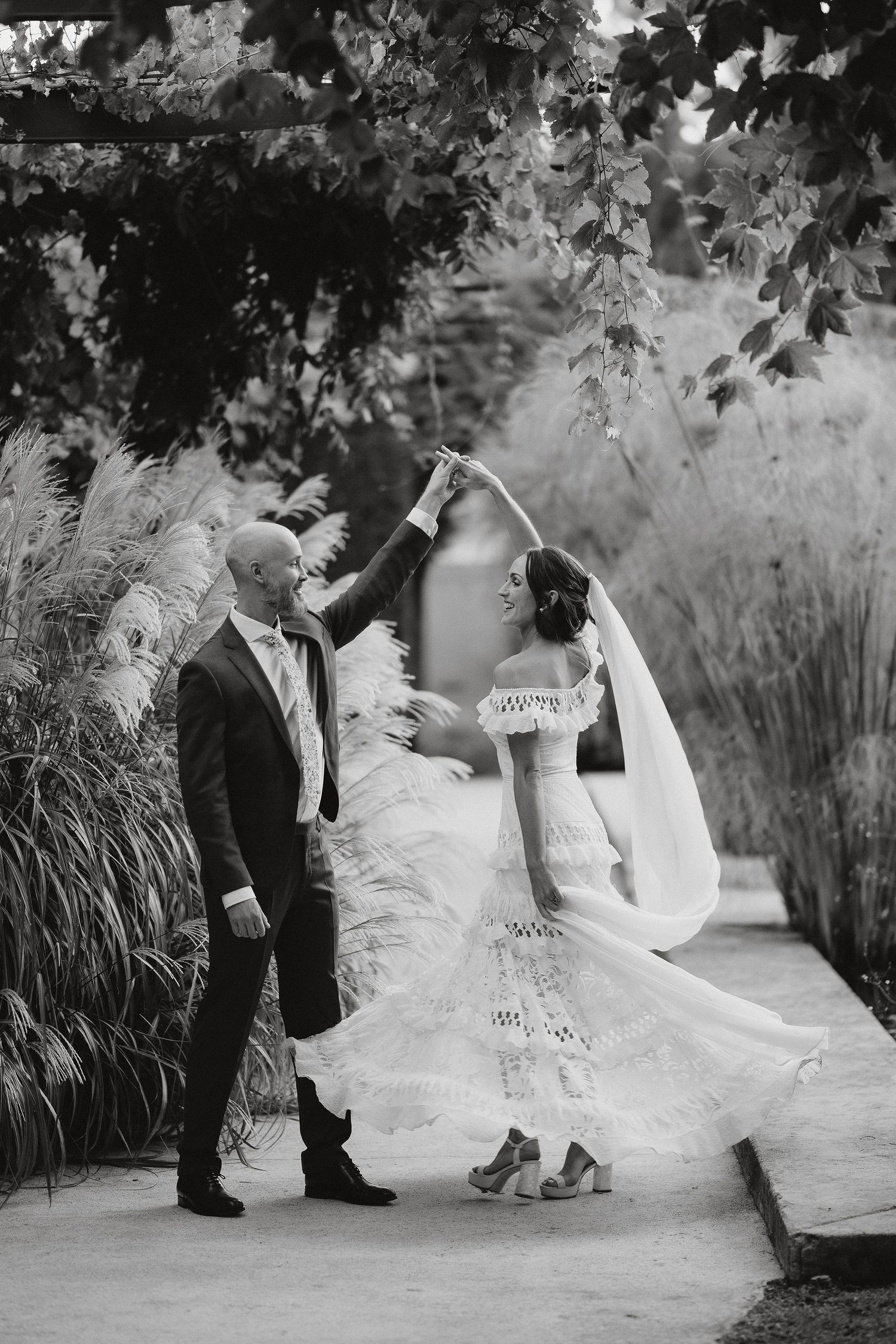 A romantic black and white wedding photograph showing a couple twirling on a garden path beneath overhanging leaves.