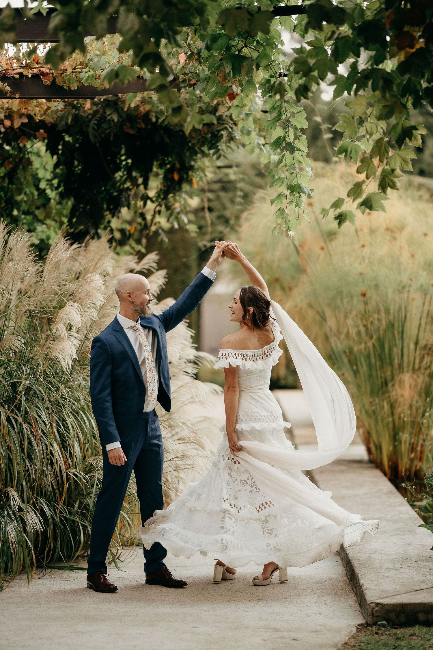 A romantic wedding moment captured outdoors as a couple dances beneath hanging foliage and tall grasses.