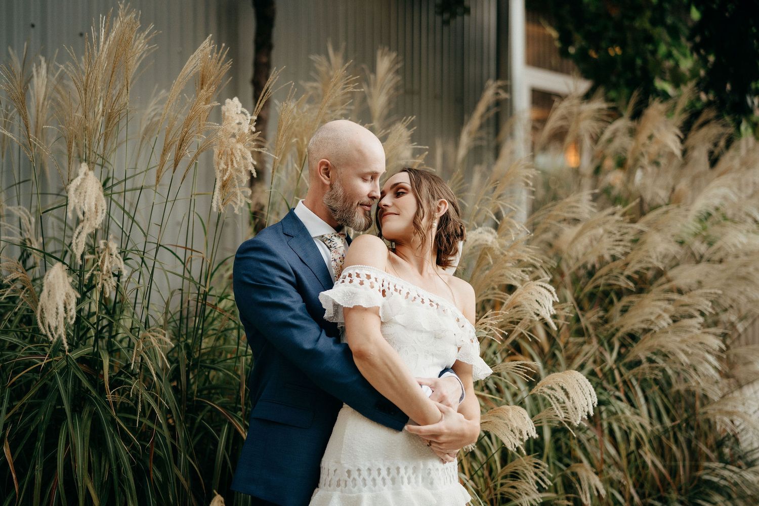 Newlywed couple embraces lovingly among tall pampas grass on their wedding day.