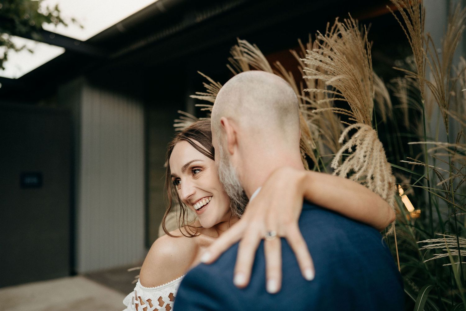 Romantic outdoor wedding moment featuring couple embracing near pampas grass against dark building.