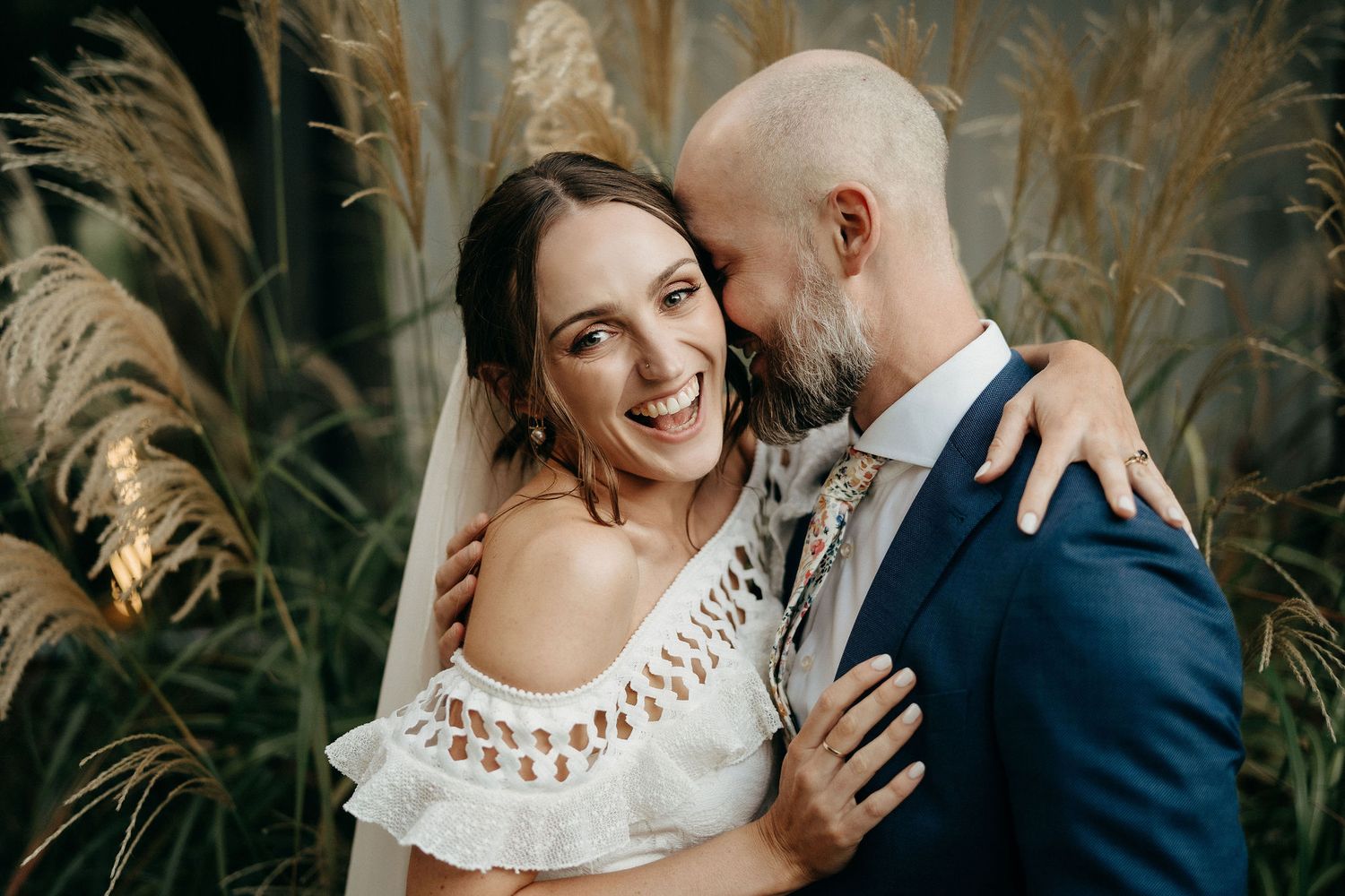 A couple embraces and smiles together in a romantic outdoor setting with pampas grass in the background.