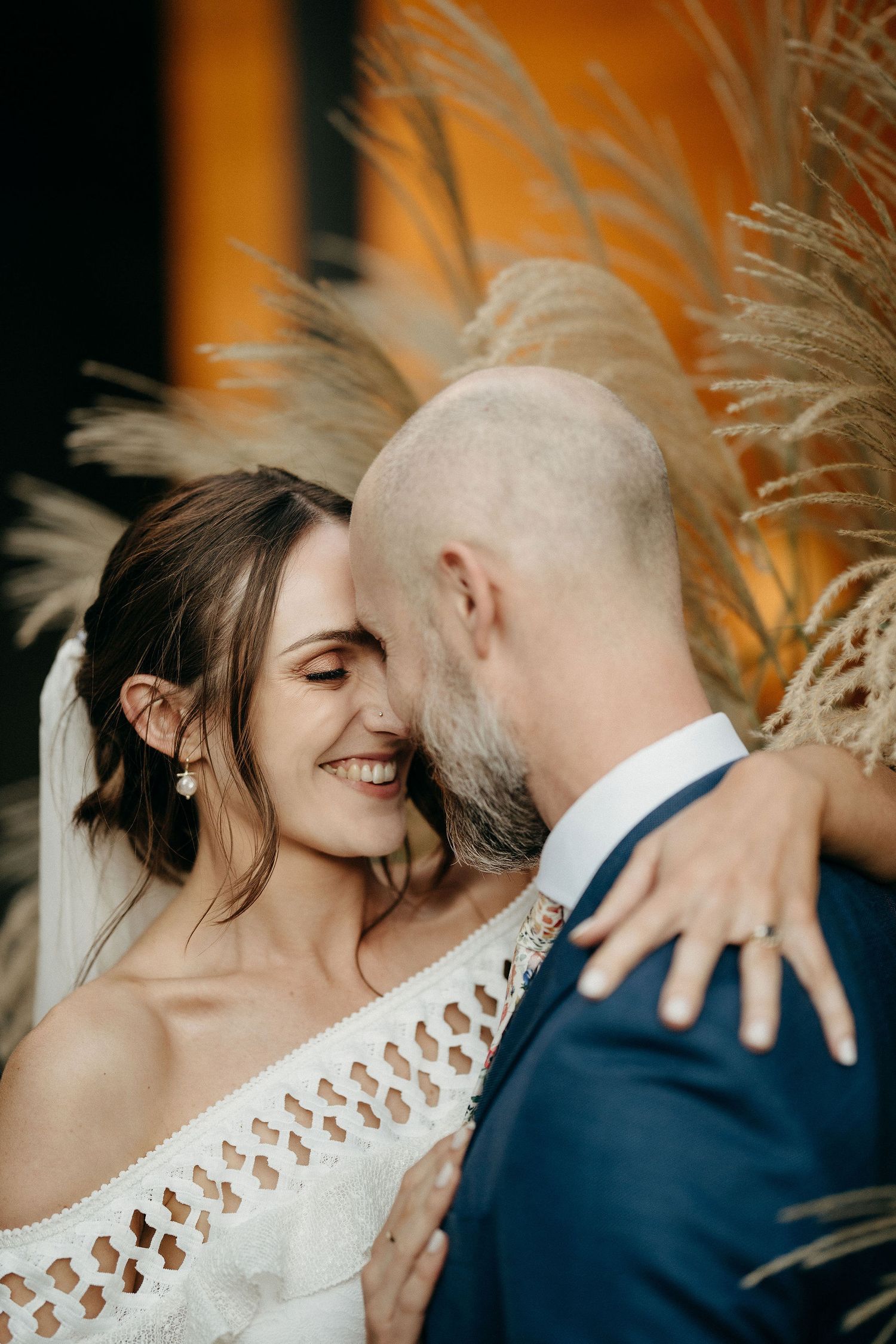 A romantic wedding moment captured against a backdrop of pampas grass with warm orange lighting.