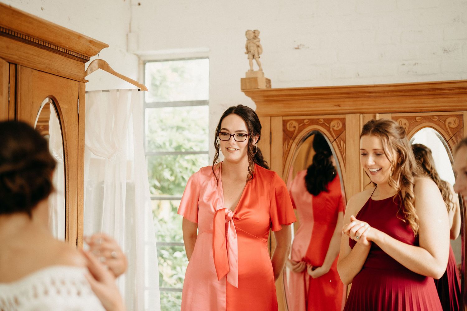 Two bridesmaids in red dresses help prepare for a wedding in a sunlit dressing room.