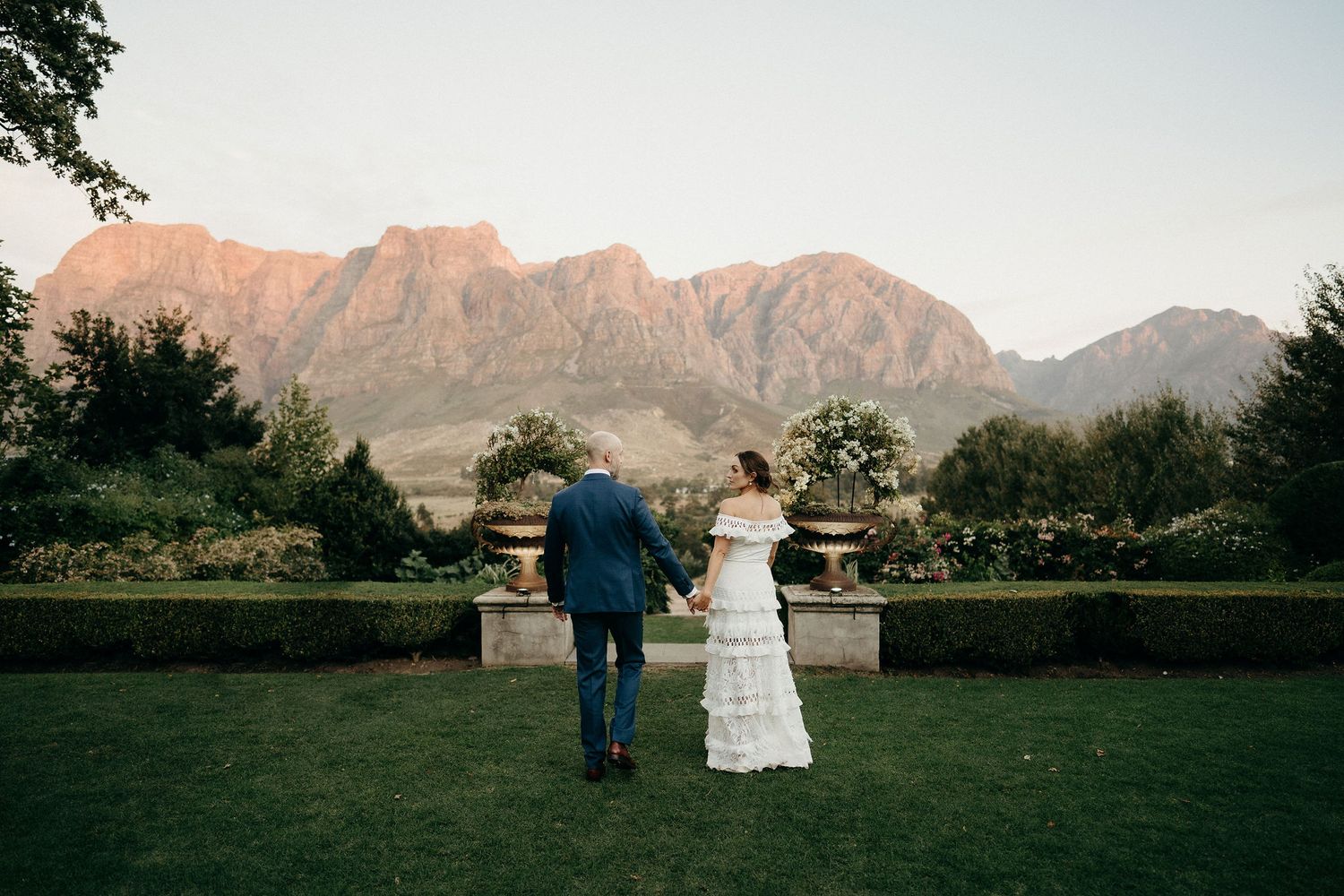 A couple gazes at majestic mountains from a formal garden terrace with stone balustrades at sunset.