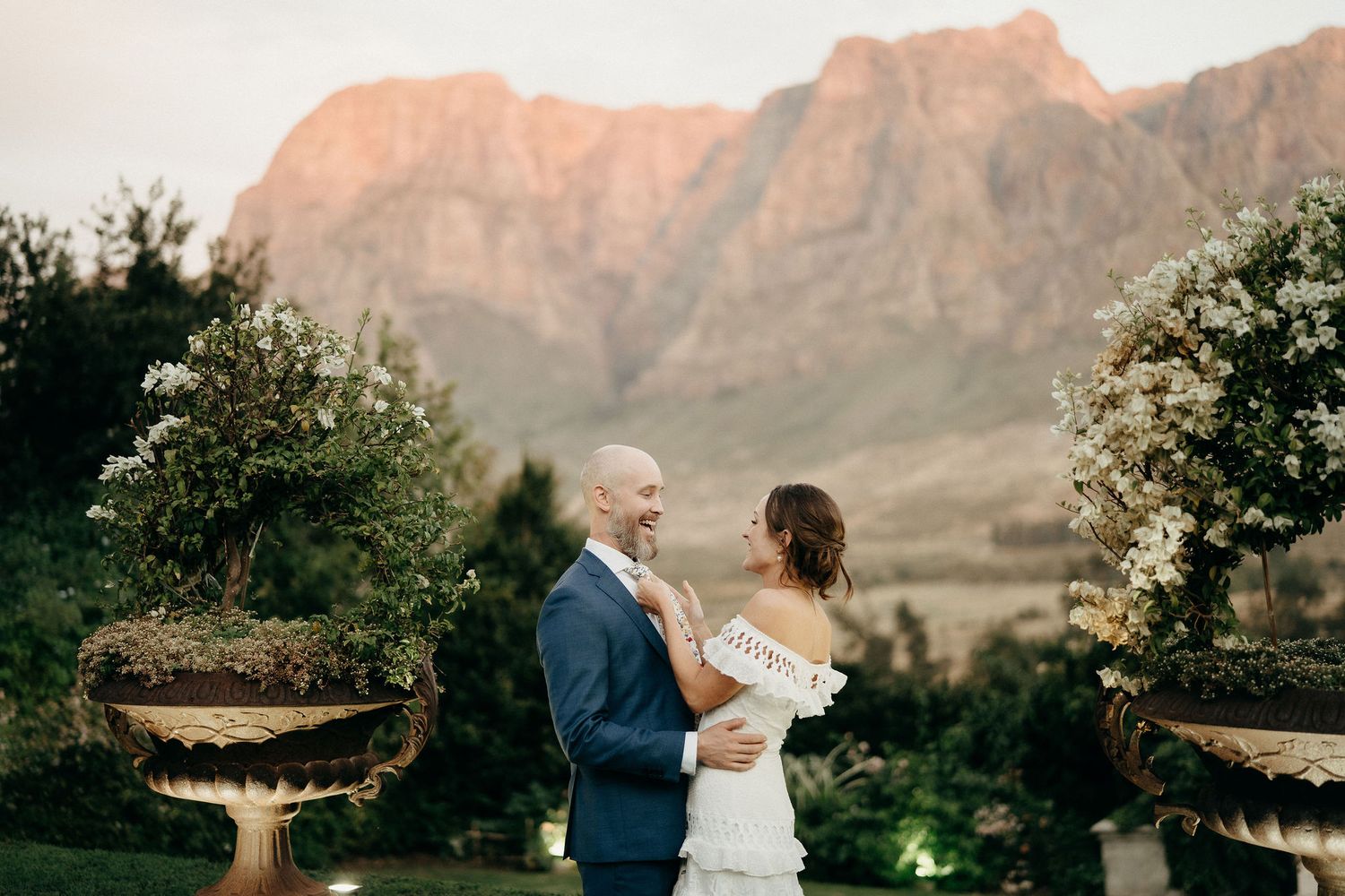 A couple shares an intimate moment at sunset with dramatic mountain peaks glowing orange in the background at a garden venue.