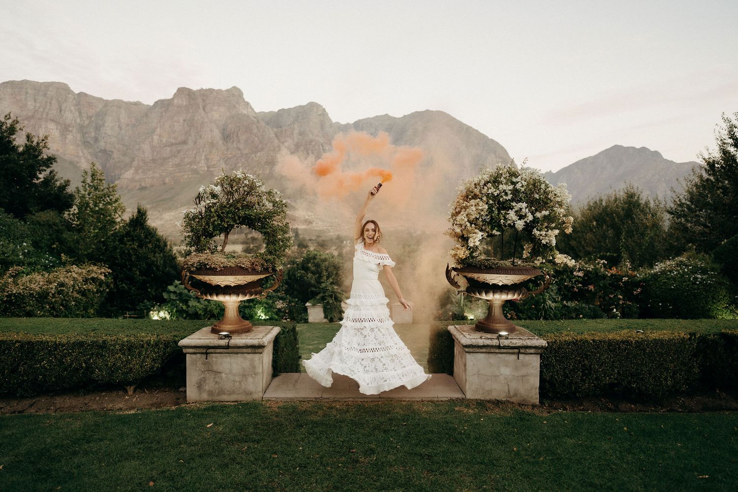 An elegant bridal photo at a garden estate with dramatic mountains in the background and ornate stone urns.