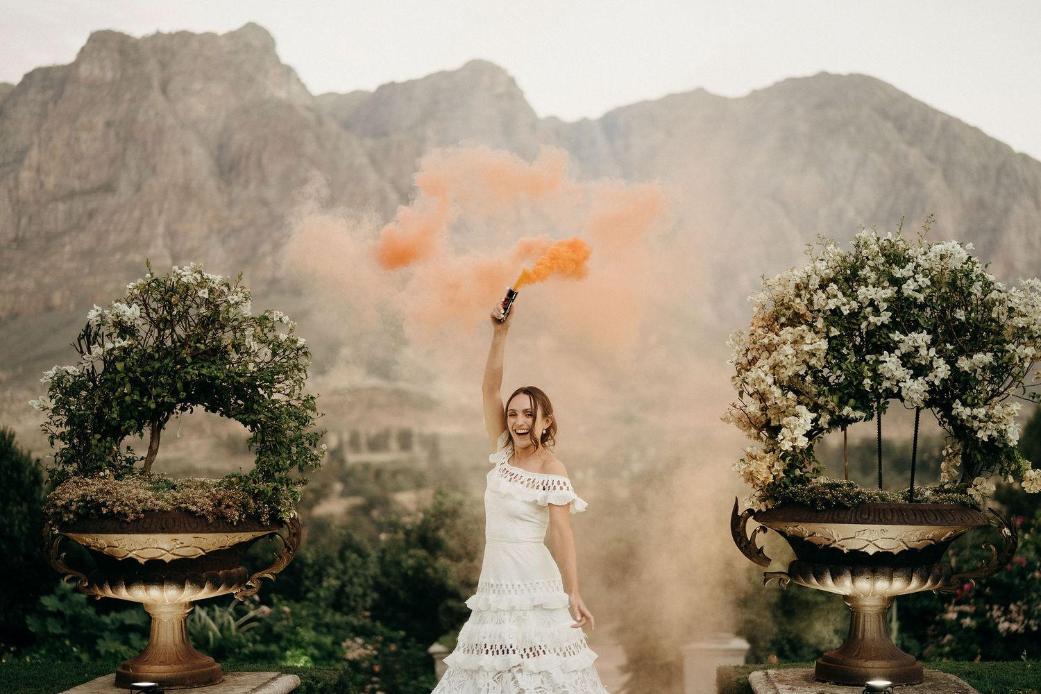 A bride in white holds an orange smoke flare against a backdrop of dramatic mountain cliffs and ornate garden urns.