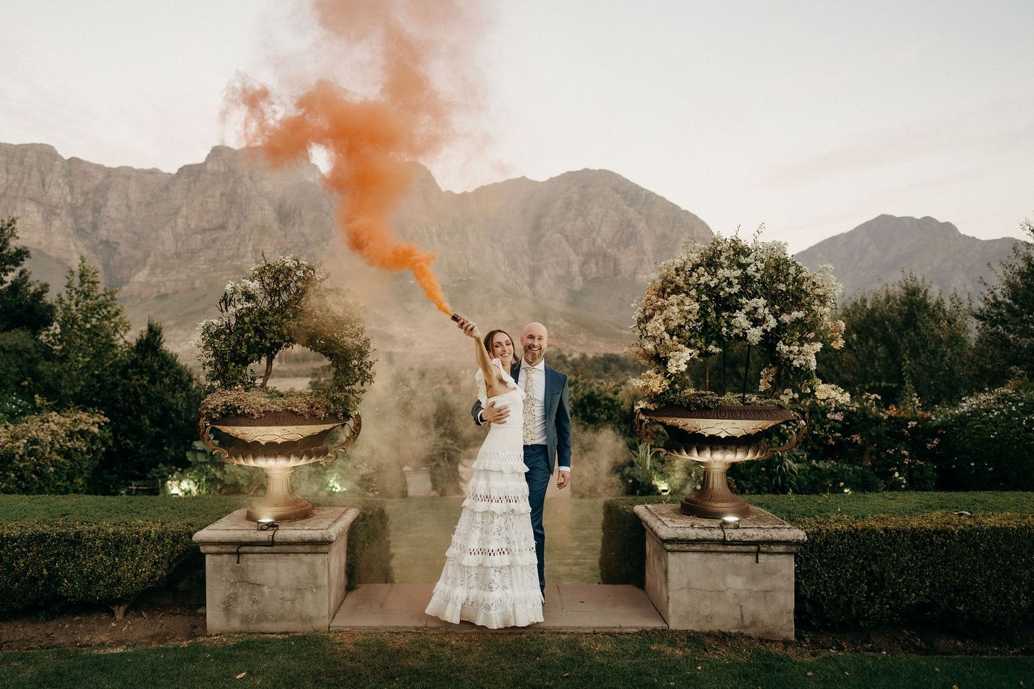 Wedding couple with orange smoke bomb in front of majestic mountain backdrop and stone pillars at outdoor venue.