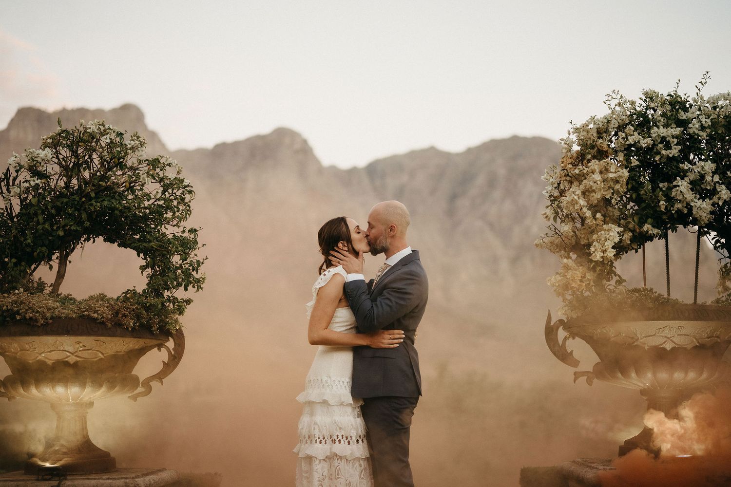 Newlyweds share a romantic moment amid swirling orange smoke with mountains and decorative urns framing the scene.