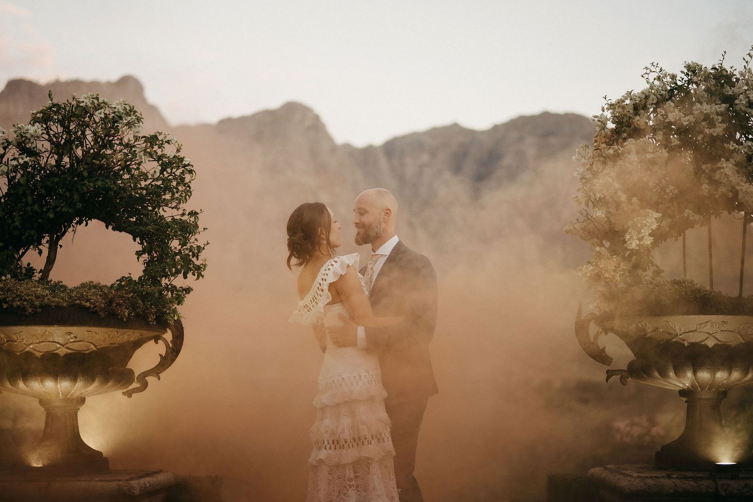 A romantic couple embraces in golden sunlight with elegant garden urns and ethereal mist surrounding them.
