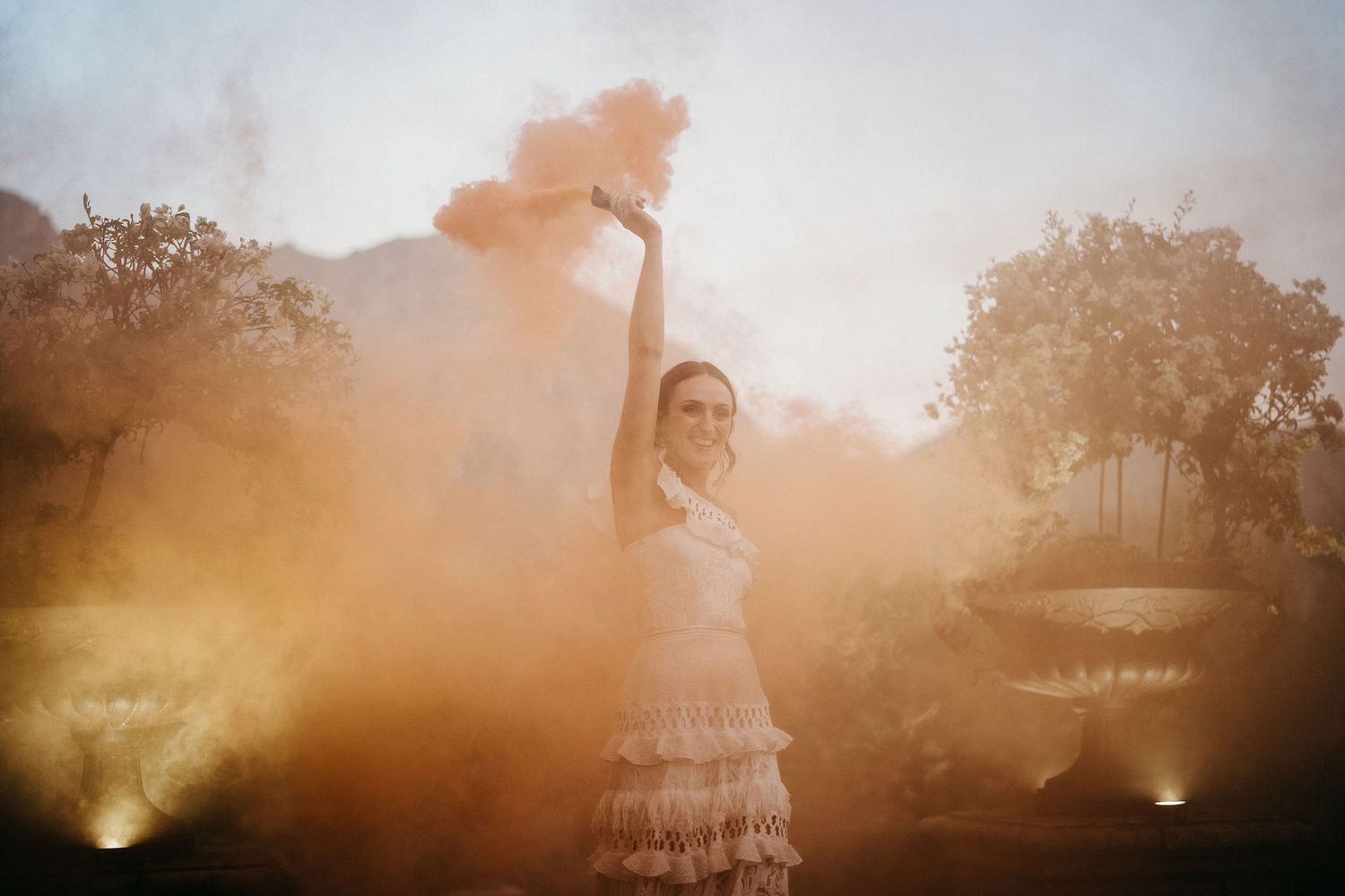 A figure in a white ruffled dress dances outdoors at sunset as golden light streams through smoke or haze in the air.